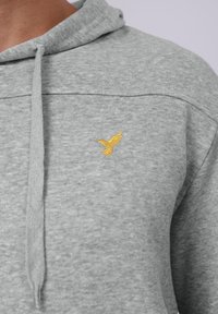 Sudadera gris con cordón, hecha de tela suave. Presenta un pequeño logotipo de un pájaro amarillo bordado en la parte izquierda del pecho.