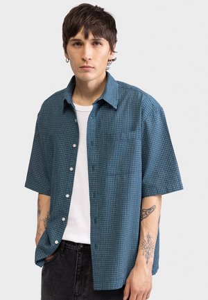 Νέος άνδρας με κοντά σκούρα μαλλιά, φοράει ασημένια σκουλαρίκια κρίκους, μπλε καρό κοντομάνικο πουκάμισο πάνω από λευκό αμάνικο και μαύρο παντελόνι.