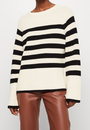 Personne portant un pull en maille blanc avec des rayures horizontales noires et un pantalon en cuir marron, visible du cou aux cuisses.
