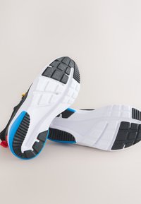 Paire de chaussures de sport noires avec semelles blanches présentant des motifs de gomme noirs et bleus, placées de manière à montrer la semelle.