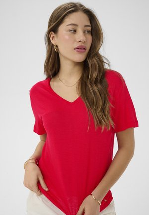 Femme aux longs cheveux bruns portant des boucles d'oreilles créoles en or et des bracelets, vêtue d'un t-shirt rouge à col en V et d'un pantalon clair, regardant sur le côté.
