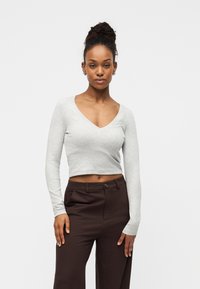 Γυναίκα με σκουρόχρωμα πλεκτά μαλλιά σε κότσο φοράει ανοιχτό γκρι cropped τοπ με μακριά μανίκια και σκουρόκαφέ ψηλόμεσο παντελόνι, στέκεται μπροστά σε λευκό φόντο.