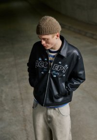 Mladá osoba nosící hnědou pletenou čepici, černou koženou bundu s nápisem „ACCEPT The Rules“, proužkované tričko a světle zbarvené kalhoty, dívající se dolů.