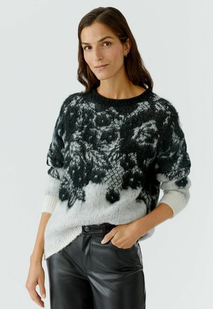 Schwarzer und weiß gemusterter Pullover mit Rundhalsausschnitt, oversized Passform und strukturiertem Strick. Getragen mit schwarzen Lederhosen, was einen lässigen Look zeigt.