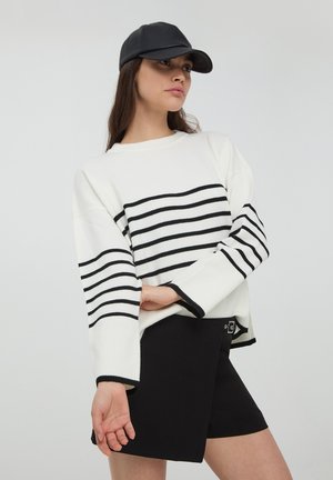 Maglione bianco con strisce orizzontali nere, texture a coste, maniche larghe. Abbinato a pantaloni corti neri con dettaglio a bottone. Cappellino nero.