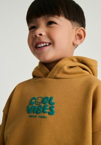 Brun hættetrøje med en blød tekstur, der har "COOL VIBES" i teal og et smilende ansigt. Designet inkluderer en stor, fastgjort hætte.