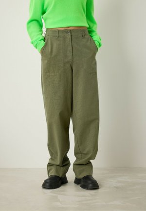 Losse olijfgroene broeken met een gestructureerd patroon, voorzien van twee voorzakken en een knoopsluiting. Gecombineerd met zwarte schoenen.