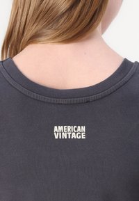 Tamno siva majica kratkih rukava s okruglim izrezom; ima natpis "AMERICAN VINTAGE" ispisan bijelo na leđima blizu ovratnika. Mekan materijal.