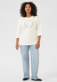 Camisa blanca de manga larga con un diseño gráfico circular en el centro, combinada con vaqueros de pierna ancha de color azul claro y zapatos planos negros.
