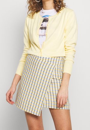 Femme portant un cardigan boutonné jaune pâle sur un t-shirt graphique et une mini-jupe asymétrique à carreaux jaunes et bleus.