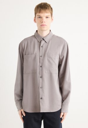 Chemise à manches longues en tissu gris doux avec un col à boutons, deux poches poitrine et une coupe droite. Présente des détails de couture subtils.