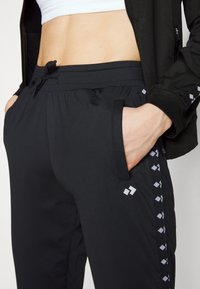 Gros plan d'une personne portant un pantalon de survêtement noir avec des bandes latérales à logo, un crop top blanc et une veste noire, la main dans la poche.
