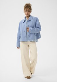 Világoskék, gyapjúból készült rövidített dzseki, szórt gallérral, elülső gombokkal és oldalsó zsebekkel; krém színű, bő szárú nadrággal párosítva.