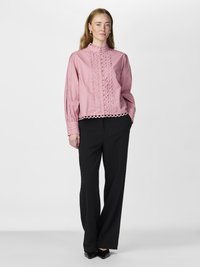 Pink blouse with intricate lace detailing along sleeves and hem, featuring a high neckline. Styled with black trousers and black shoes.