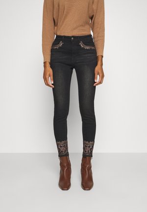 Donna che indossa jeans aderenti neri con motivi floreali ricamati sulle tasche e sui polsini, abbinati a stivaletti marroni e a un maglione color senape.