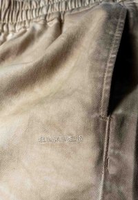 Shorts en coton beige avec une ceinture élastique, dotés d'une poche latérale et d'un lettrage discret "ELIAS RUMELIS" brodé sur le tissu.