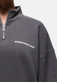 Felpa grigia in tessuto morbido con colletto alto e zip a metà. Presenta la scritta bianca "GOODFOR NOTHING" sul petto.