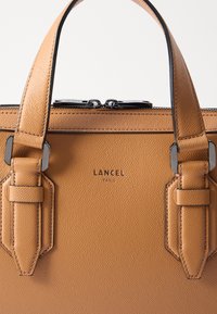 Borsa a mano in pelle in tonalità cuoio con superficie testurizzata, doppi manici superiori e dettagli in metallo. Presenta il nome del marchio impresso "Lancel Paris".