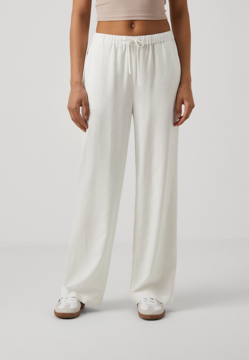Białe, lekkie, luźne spodnie z elastycznym paskiem w talii i sznurkiem do regulacji. Gładka struktura, szerokie nogawki, w zestawieniu z białymi sneakersami.