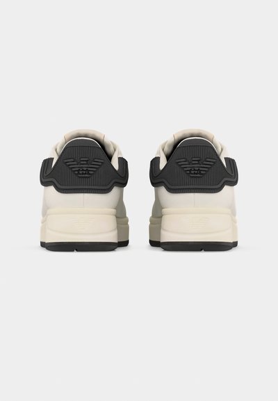 Ζευγάρι λευκών αθλητικών παπουτσιών με μαύρες φτέρνες που φέρουν ανάγλυφο λογότυπο GA, εμφανιζόμενα από πίσω σε απλό ανοιχτό φόντο.