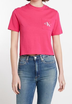 Nainen, joka pukeutunut kirkkaan pinkkiin lyhyeen T-paitaan, jossa on pieni Calvin Klein -logo rintataskussa, ja korkeavyötäröisiin sinisiin farkkuihin.