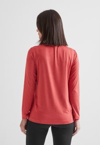 Woman with shoulder-length brown hair wearing a long-sleeve red top and black pants, facing away against a plain background.