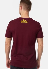 Burgundi színű, pamut póló kerek nyakkal, sárga logónyomattal a háton. Rövid ujjú és ellazult fazonú.