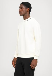 White crewneck sweatshirt with ribbed cuffs and hem, featuring a logo detail on the shoulder. Material appears soft and textured.