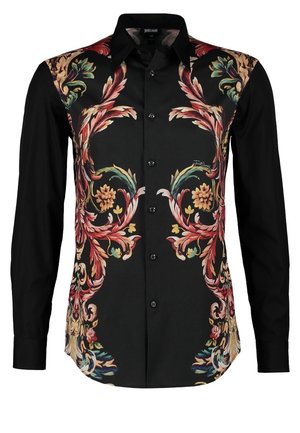 Persona que lleva pantalones negros ajustados, camisa negra con estampado floral abotonada, zapatos de vestir negros, y lleva una mochila negra sobre un hombro.