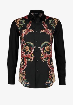 Persona que lleva pantalones negros ajustados, camisa negra con estampado floral abotonada, zapatos de vestir negros, y lleva una mochila negra sobre un hombro.