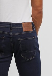 Tumši zili džinsi ar gaiši brūnu ādas uzlīmi aizmugurējā jostas daļā, redzamām vīlēm un klasisku četru kabatu dizainu.