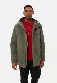 Groene waterdichte jas met capuchon, twee voorzakken en trekkoorden, gedragen over een rood shirt met een bergafbeelding.