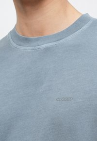 Gros plan sur une personne portant un t-shirt bleu clair à col rond avec un texte en relief subtil "CLOSED" sur la poitrine.