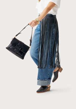 Femme portant un jean bleu avec une ceinture à franges en cuir noir, un haut blanc, des mules tissées noires à talons, et tenant un sac à main en cuir noir.