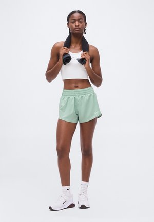 Femme athlétique portant un crop top blanc, un short vert menthe, des chaussettes blanches et des baskets, tenant une serviette noire autour du cou.