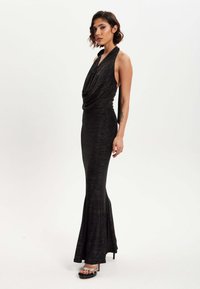 Black, sleeveless dress with draped neckline, fitted silhouette, flared hem, and subtle shimmer. Paired with strappy high-heeled sandals.