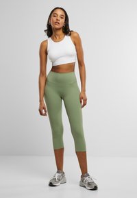 Hvit avkortet tanktopp og olivengrønne høytliv leggings, laget av elastisk materiale, kombinert med grå joggesko.