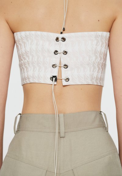 Top corto bianco con motivo grafico, spalle regolabili con lacci e occhielli in argento, abbinato a pantaloni beige chiaro a vita alta.