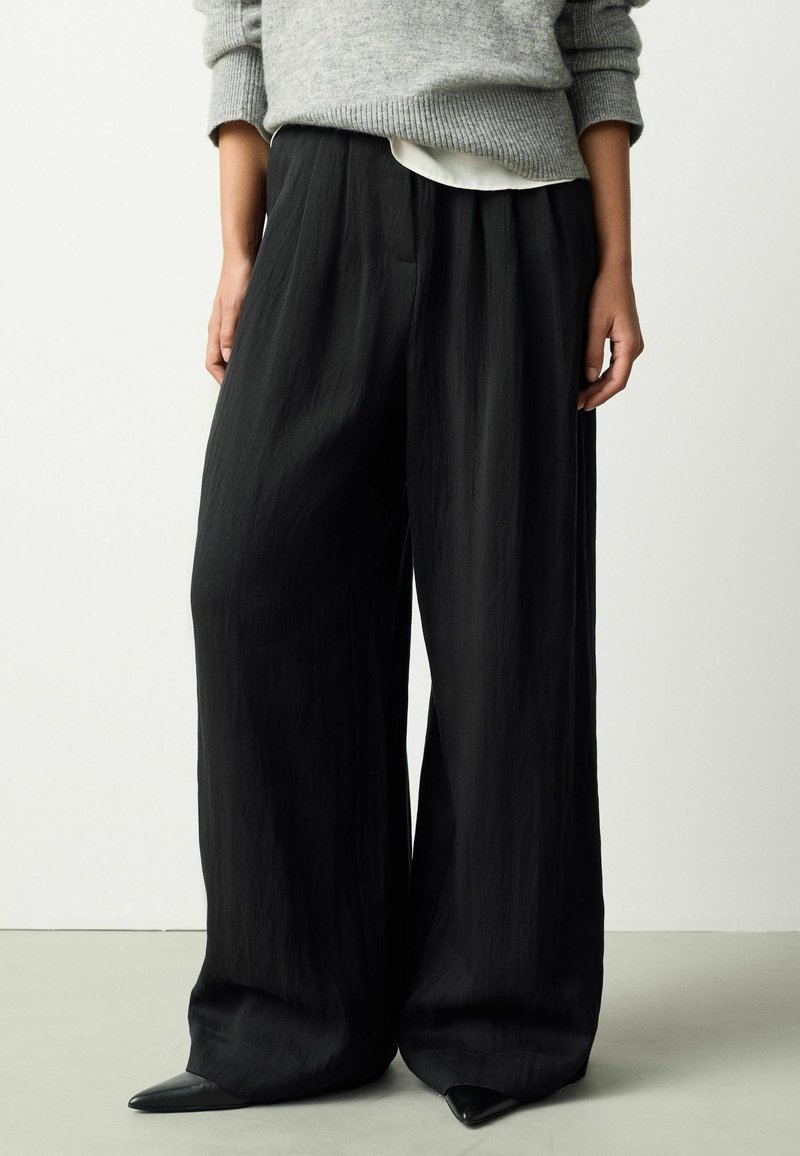 Svarta vida byxor i ett lätt, strukturerat tyg. Har en flödande design och små veck för extra volym.