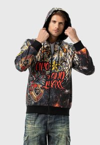 Reißverschluss-Hoodie mit lebendigem Grafikdesign, das einen Adler, Rüstungsschilde und fett gedruckte rote Schrift zeigt. Hergestellt aus weichem Stoff; schwarze gerippte Manschetten.