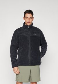 Veste en polaire noire avec un col haut, fermeture éclair intégrale, poches latérales et un logo discret sur la poitrine. Tissu doux et texturé.