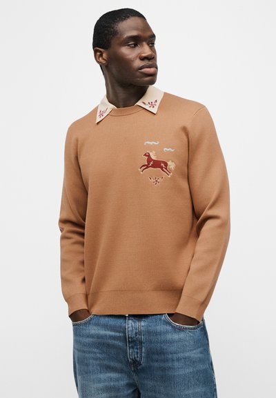 Άνδρας που φοράει μπεζ πουλόβερ με κέντημα κόκκινου αλόγου και σύννεφου, λευκό γιακά με κόκκινα σχέδια και μπλε τζιν, με τα χέρια στις τσέπες.