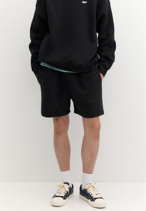 Personne portant un sweat-shirt noir et un short, des chaussettes blanches et des baskets noires et blanches, debout contre un mur clair uni.