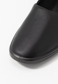 Chaussure noire à enfiler avec un dessus en simili cuir lisse, bout rond et détails de couture subtils. La semelle a un léger bord de plateforme.