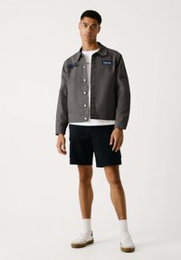 Graue Jeansjacke mit Knopfverschluss, weißen Nähten und gesticktem blauen Branding; kombiniert mit schwarzen Shorts und weißen Sneakers.