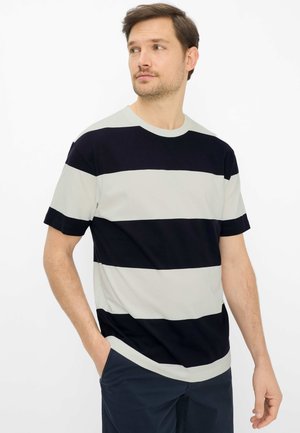 Uomo che indossa una maglietta a maniche corte a larghe strisce orizzontali bianche e nere con pantaloni scuri, in piedi con una mano in tasca, che guarda verso sinistra.