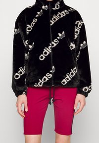 Černá bunda z umělé kožešiny na zip s opakujícím se bílým logem Adidas, s vysokým límcem a elastickými manžetami, kombinovaná s růžovými přiléhavými šortkami.