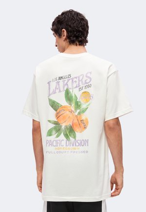 Personne portant un t-shirt blanc avec le texte Los Angeles Lakers, des ballons de basket et un graphisme de feuilles vertes dans le dos.