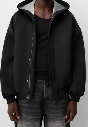 Dunkelhäutige Person, die einen schwarzen Kapuzenpullover mit Reißverschluss und grauer Innenkapuze sowie ausgewaschene schwarze Jeans trägt, steht vor einem schlichten Hintergrund.