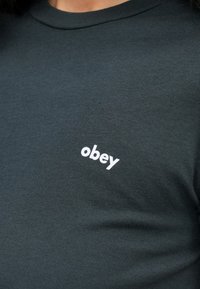 Mørkegrønn bomull T-skjorte med et lite hvitt "obey"-logo på venstre brystområde. Klassisk crewneck-design med en glatt tekstur.